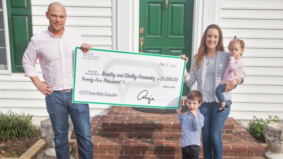 husband and wife with two small children holding big check in front of home